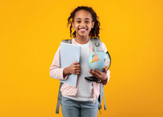 Portrait of cheerful young black girl holding Earth world globe and notebook, wearing backpack and looking at camera isolated on yellow color background at studio. Education in school concept
