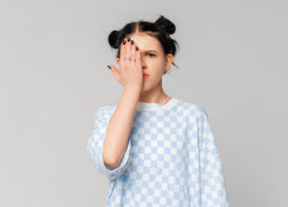 Cute dark-haired teen girl making facepalm, child covers half of face with palm, seriously looks with one eye at camera, stands in trendy t shirt over light grey background