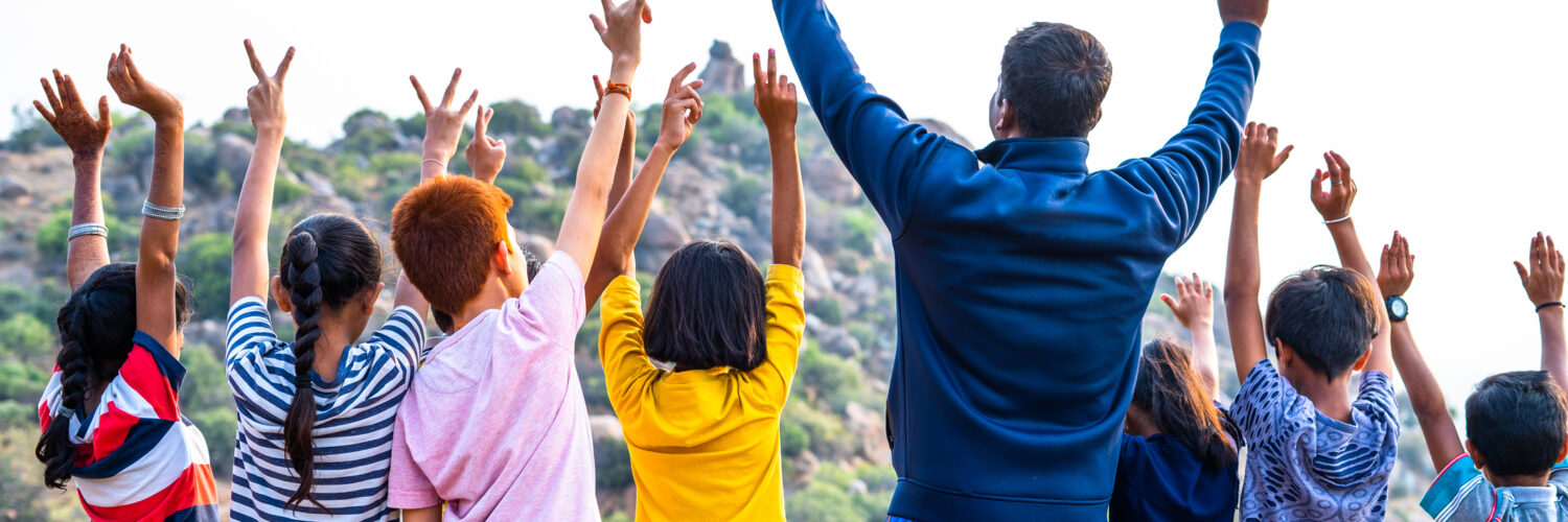 Back view shot of group of teenage kids with teacher celebrating by raising hands on top of hill after reaching destination - concept of summer holiday, trekking, outdoor adventures activities and bonding