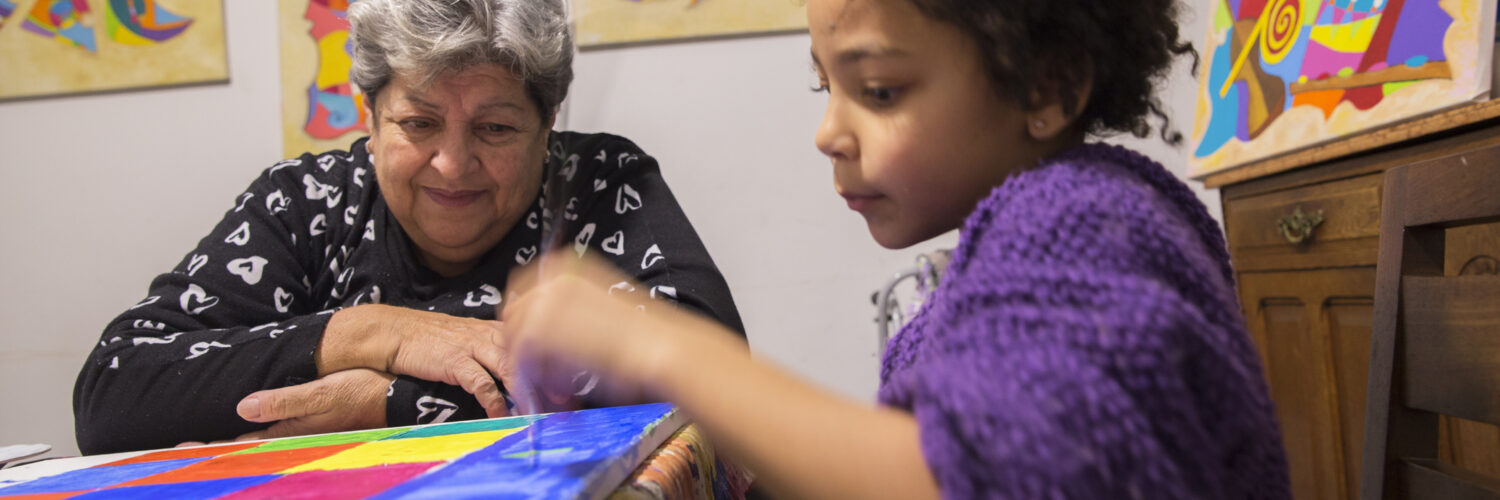 Latin grandma and cute mixed race girl painting on canvas at home.