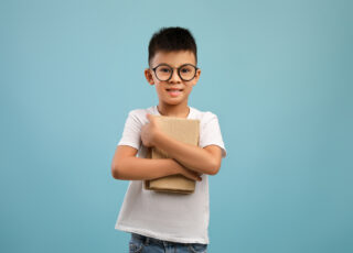 Cute little asian boy wearing eyeglasses holding stack of books and looking at camera, nerdy korean male child enjoying reading and learning, standing isolated over blue studio background, copy space