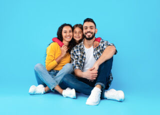 Loving Arab Family Hugging Sitting In Studio Over Blue Background. Shot Of Happy Little Daughter Embracing Her Parents Expressing Love, Smiling To Camera. Full-Length